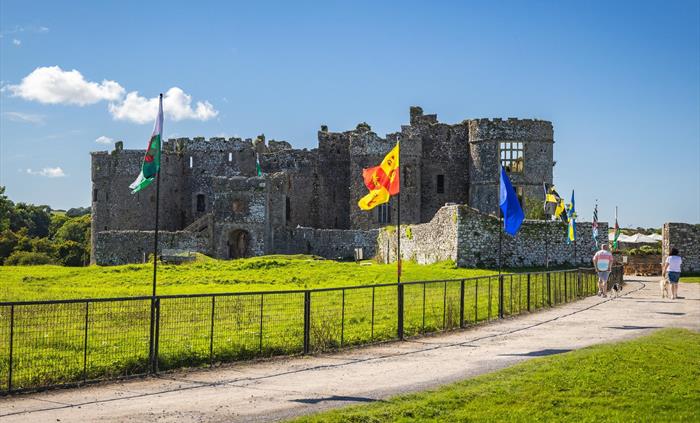 Image of Carew Castle where two dog walkers and walking up the path towards the Castle. Also several types of flags on display on fencing alongside th